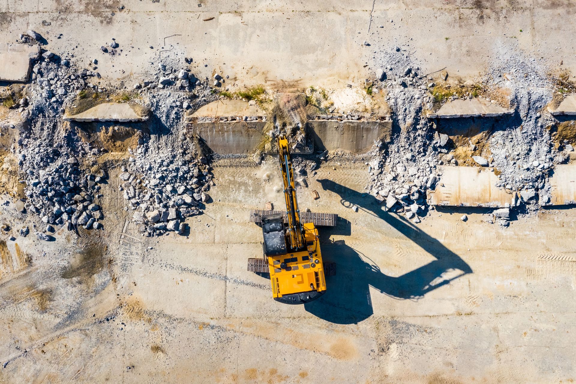 Aerial view of a yellow excavator demolishing concrete structure, piles of debris on either side, sunny day.