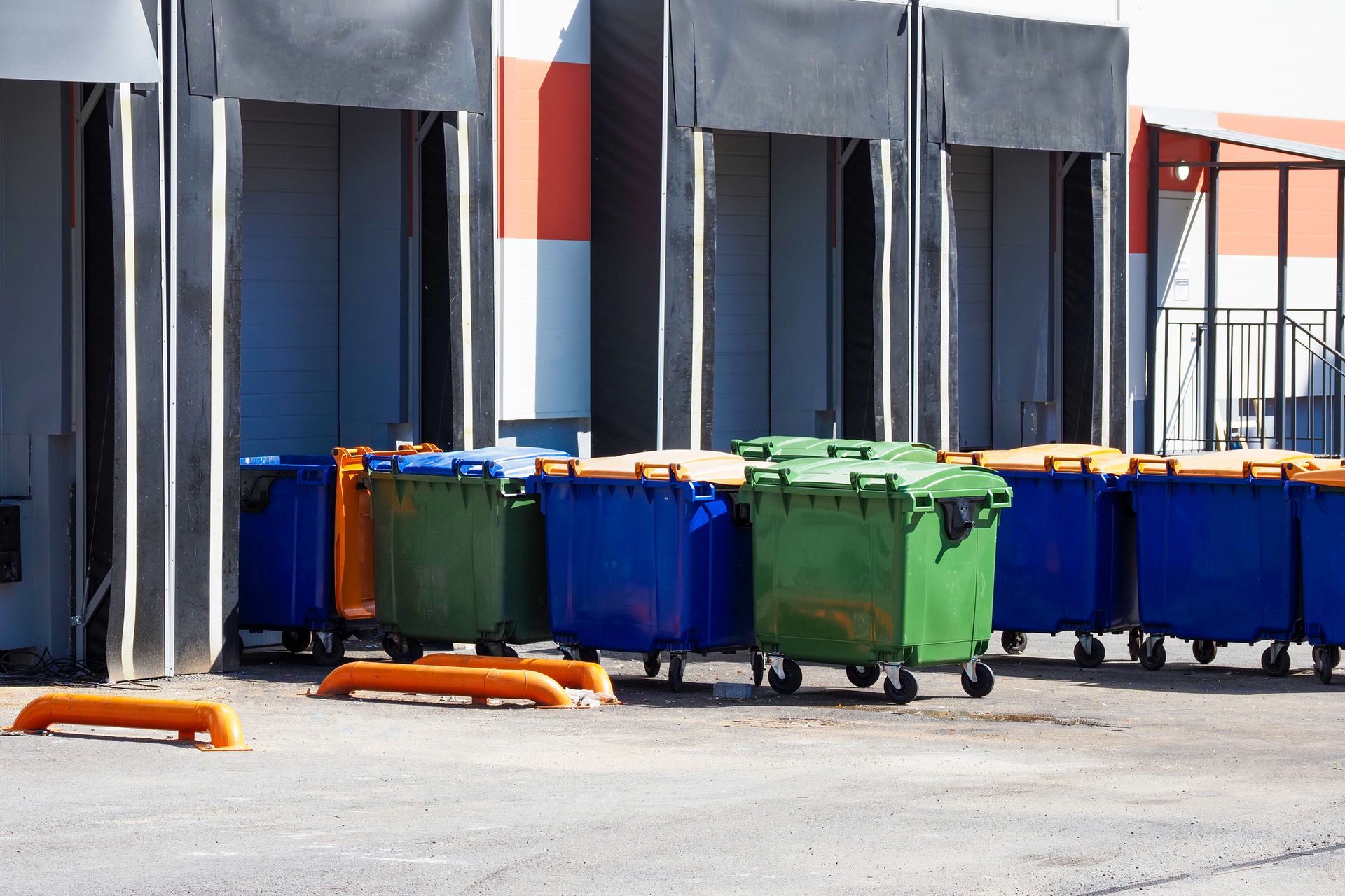 Blue and green garbage bins lined up at a loading dock with orange parking barriers.