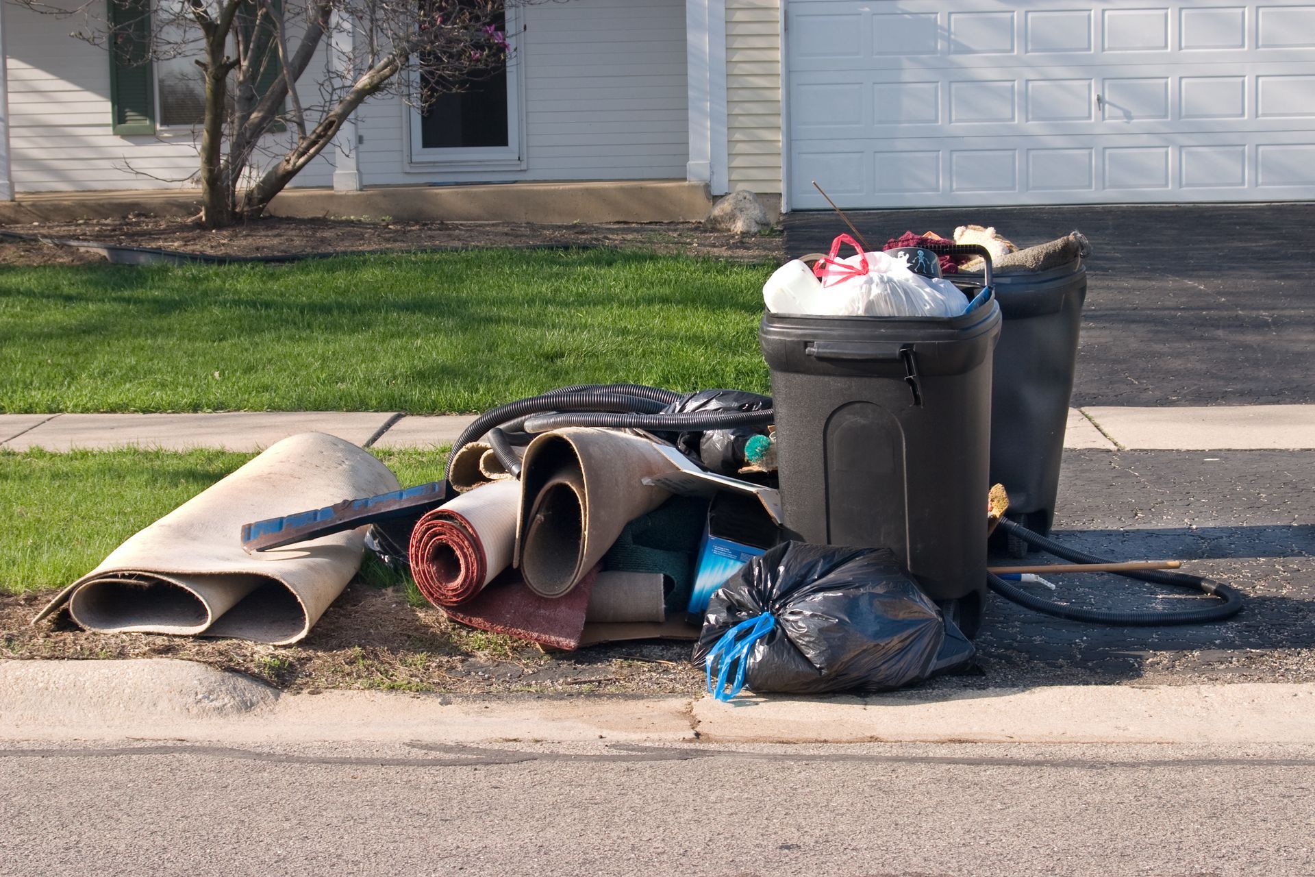 Trash and debris piled on curb: carpet rolls, trash cans, and a black bag in front of a house.