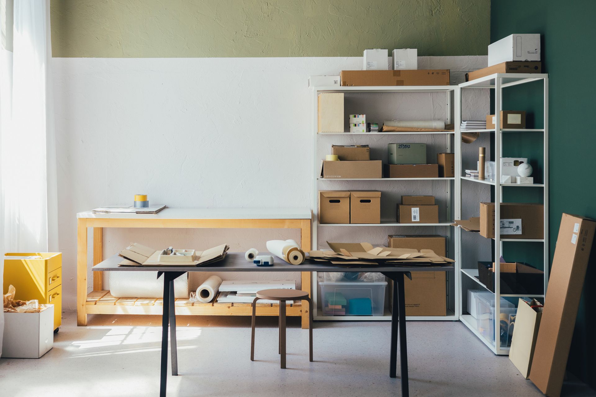 Craft room with shelving, work tables, and packing supplies. White, green, and wood-tone decor with boxes and materials.