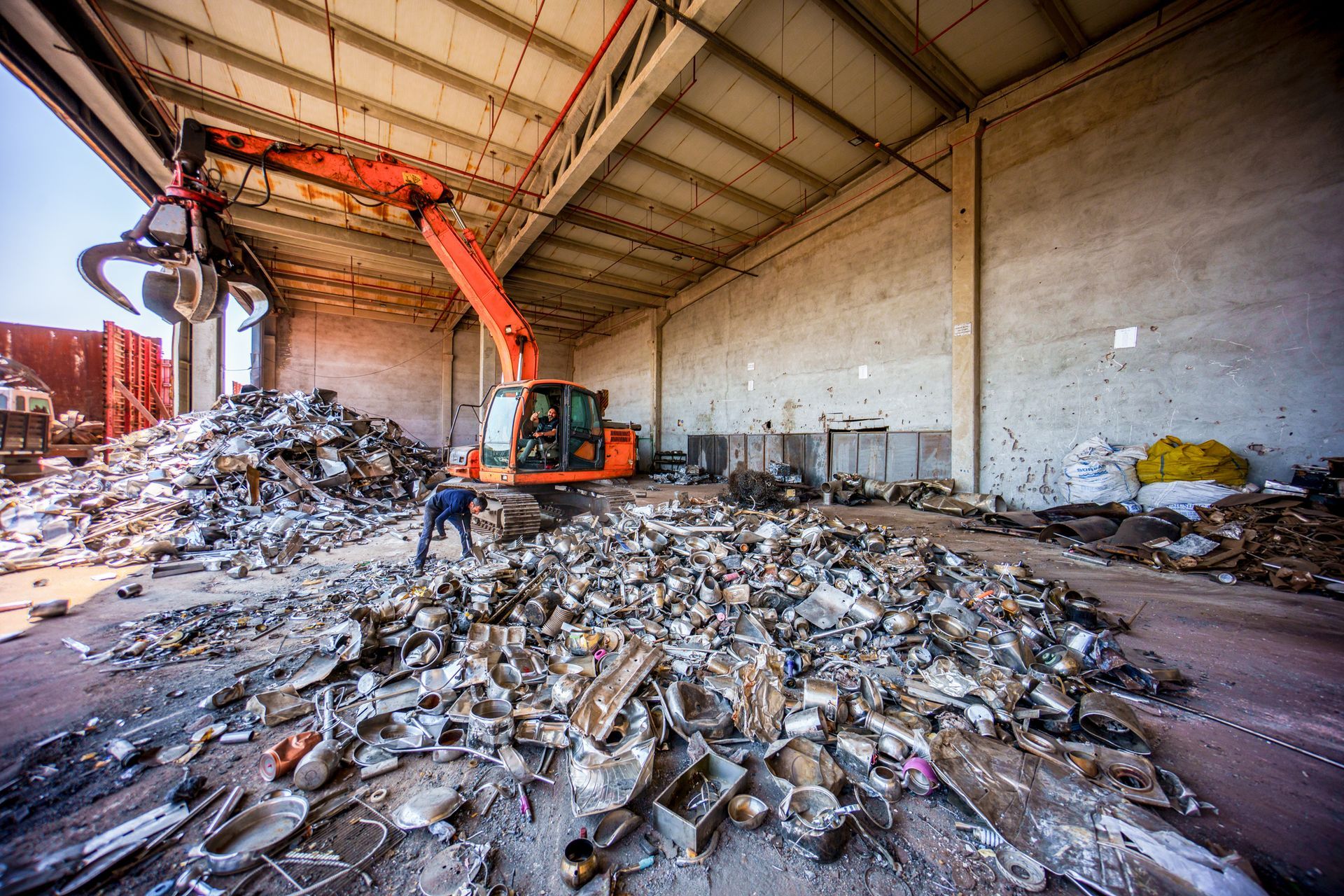 An excavator sorting scrap metal inside a warehouse; worker nearby, red and gray.