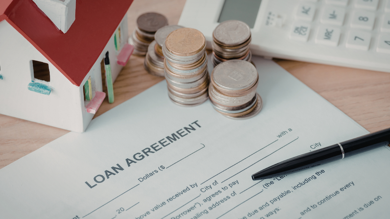 Loan agreement, toy house, coins, calculator, and pen on a wooden surface, suggesting financial concepts.