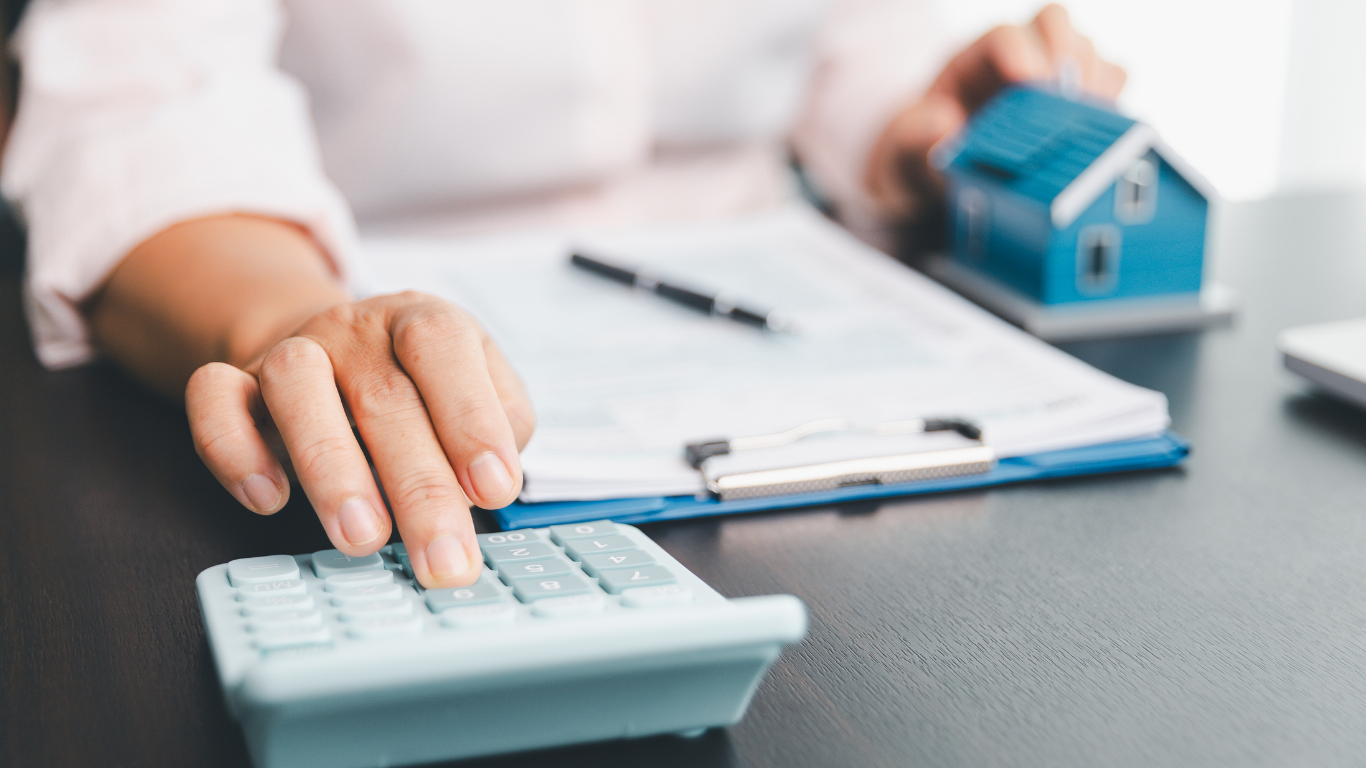 Person using calculator with document, pen, and toy house on a desk.