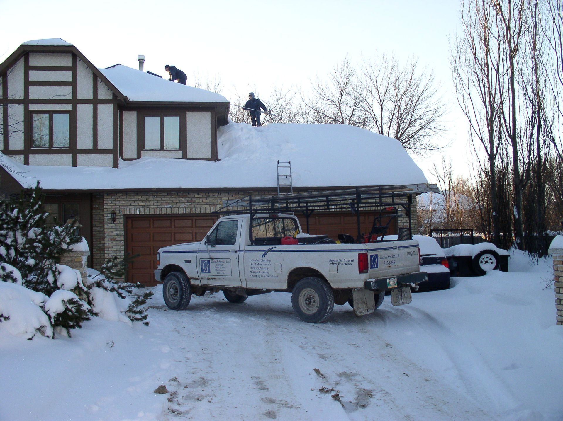 Snow-covered house with workers removing snow from the roof; a truck is parked in the driveway.