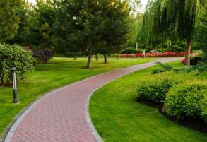 Brick pathway winds through a green park, bordered by grass, trees, and shrubs.