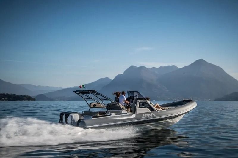 Motoscafo grigio su un lago con due persone, montagne sullo sfondo.