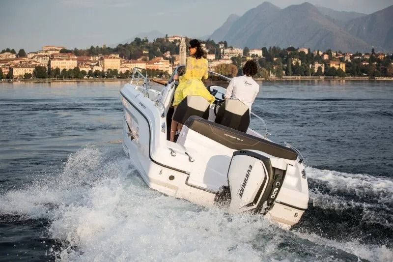 Motoscafo bianco con due persone a bordo, in navigazione sull'acqua. Città e montagne sullo sfondo.