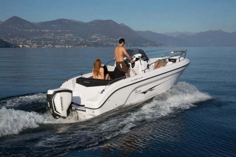 Motoscafo bianco sull'acqua con due persone, montagne sullo sfondo.