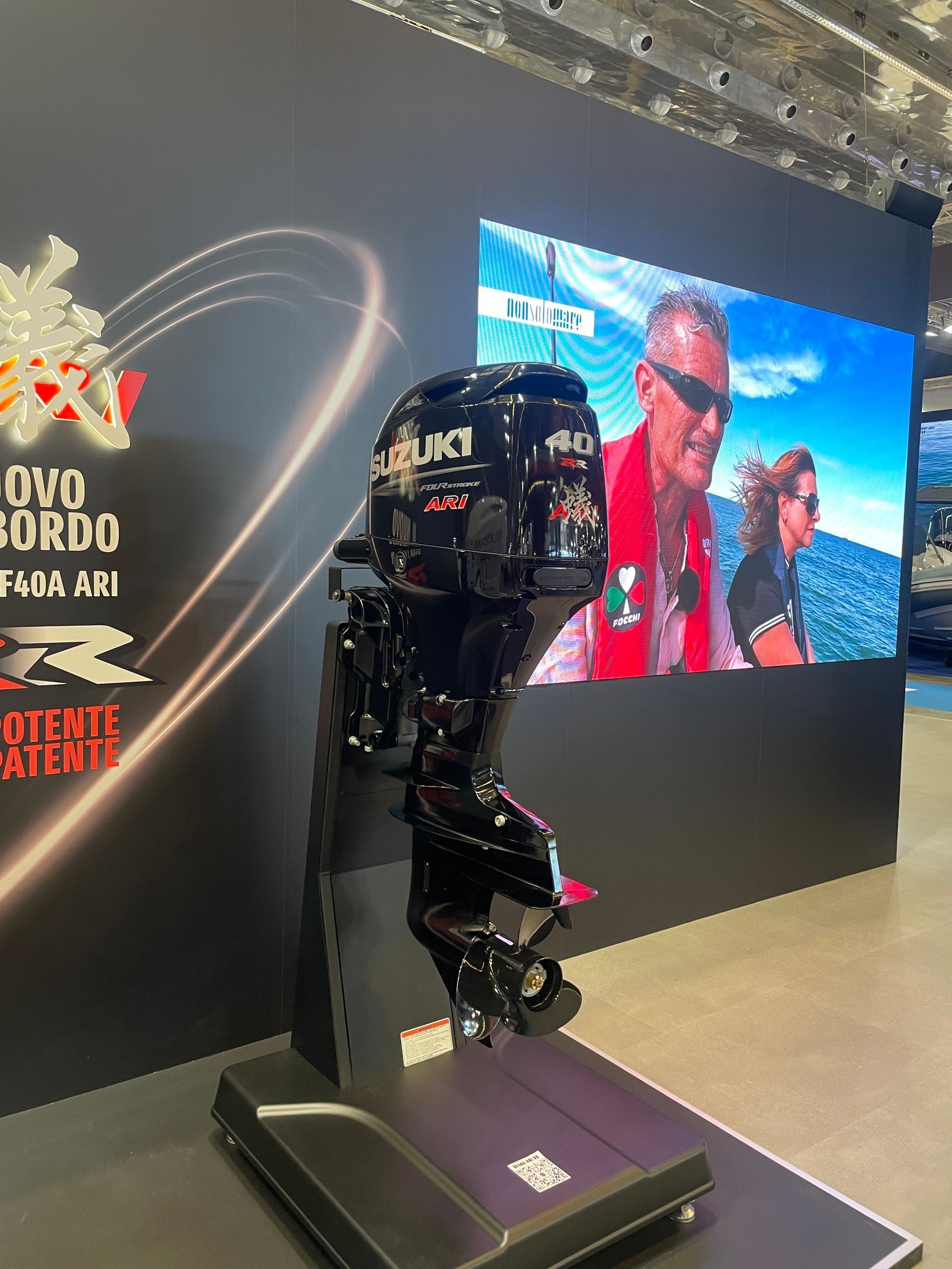 Motore fuoribordo Suzuki nero esposto a un evento. Uno schermo mostra una persona che indossa un giubbotto di salvataggio.