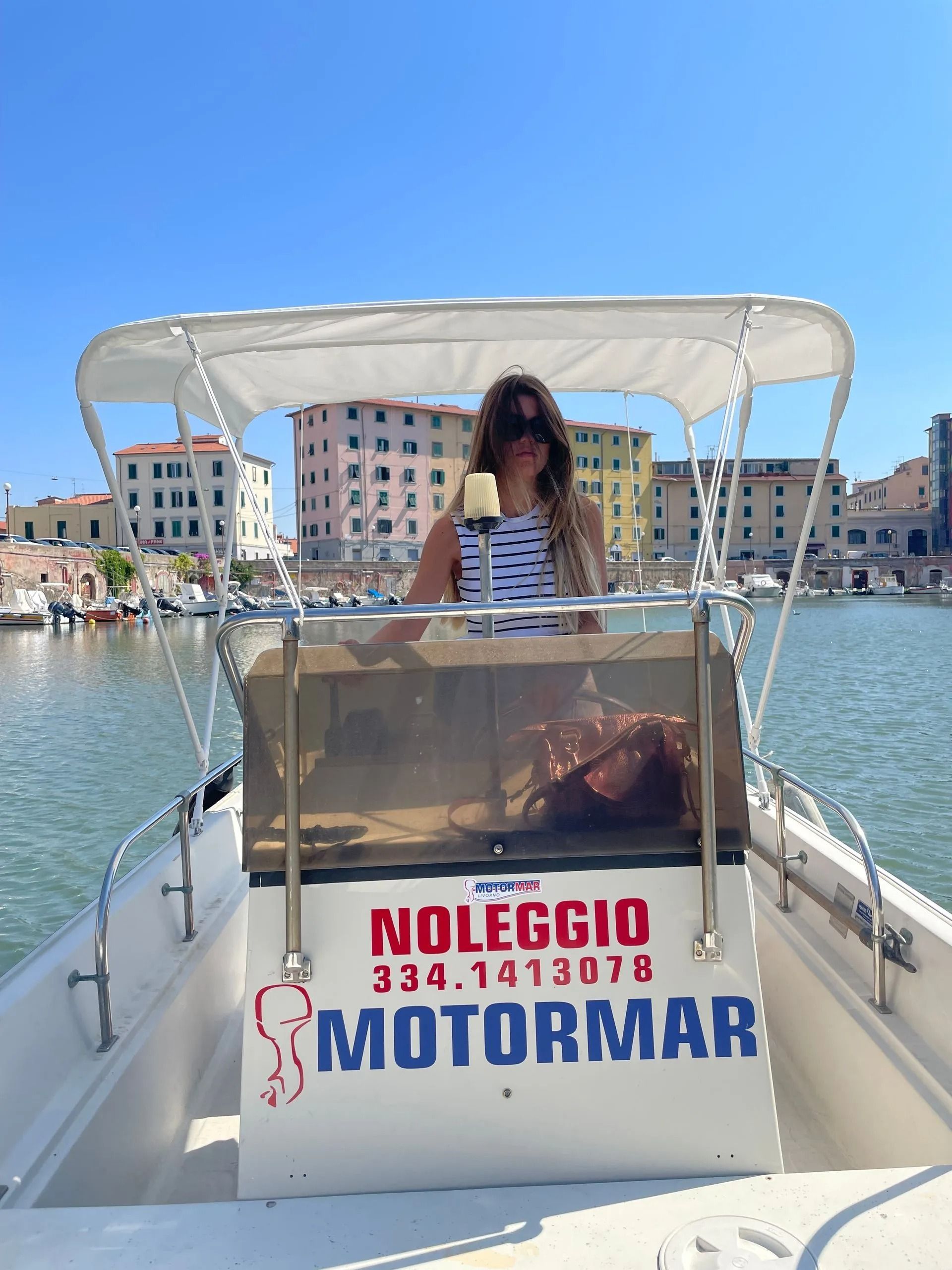 Donna su una barca, con un drink in mano. La barca è sull'acqua, con edifici cittadini sullo sfondo.