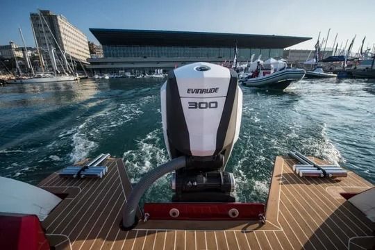 Motore fuoribordo su una barca in un porto, che crea una scia. Sullo sfondo un edificio e altre imbarcazioni.