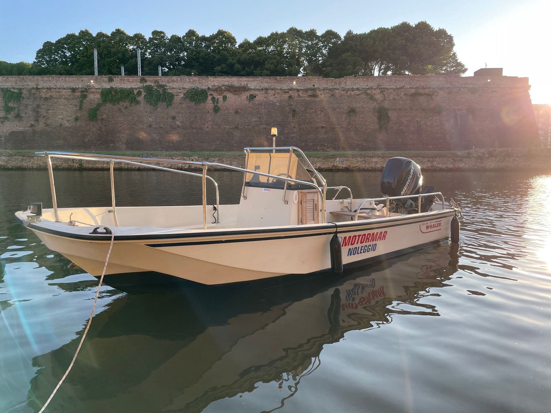 Una barca beige galleggia sull'acqua vicino a un muro di pietra al tramonto. Sul lato c'è la scritta