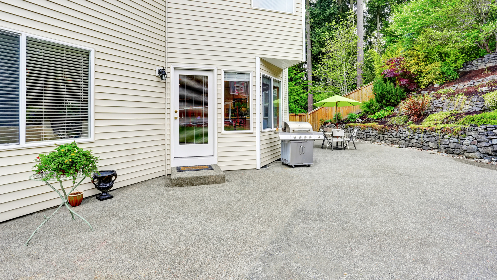 Backyard patio with light siding, a door, and a grill, overlooking a sloped garden and trees.