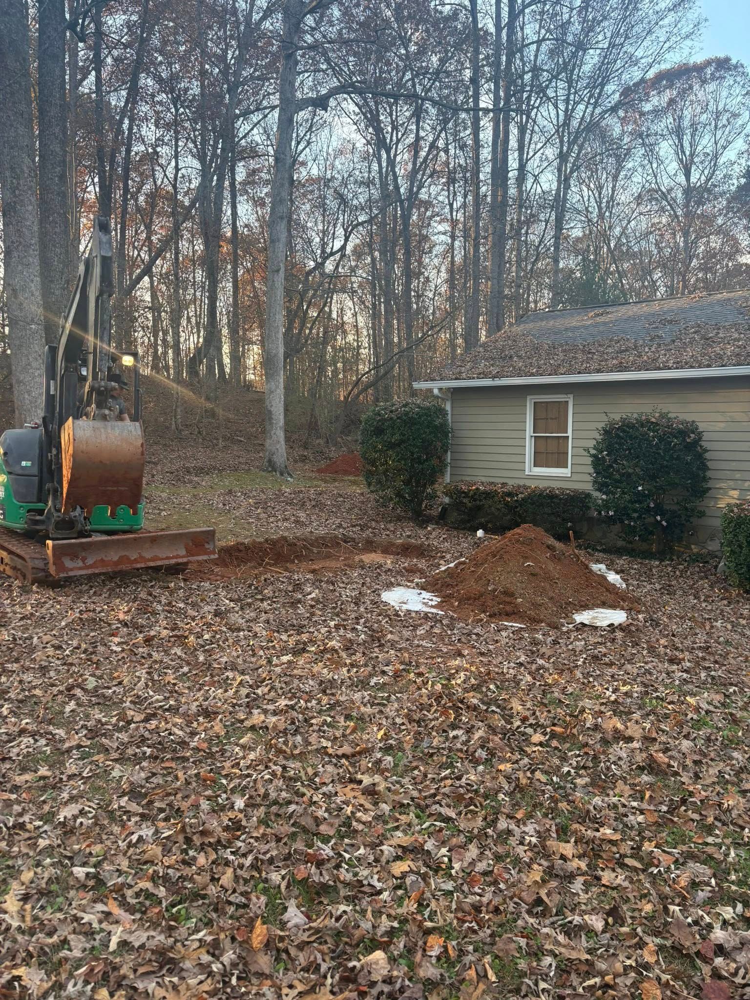 Backyard with excavator, dirt pile, and a small shed beside leaf-covered ground and trees