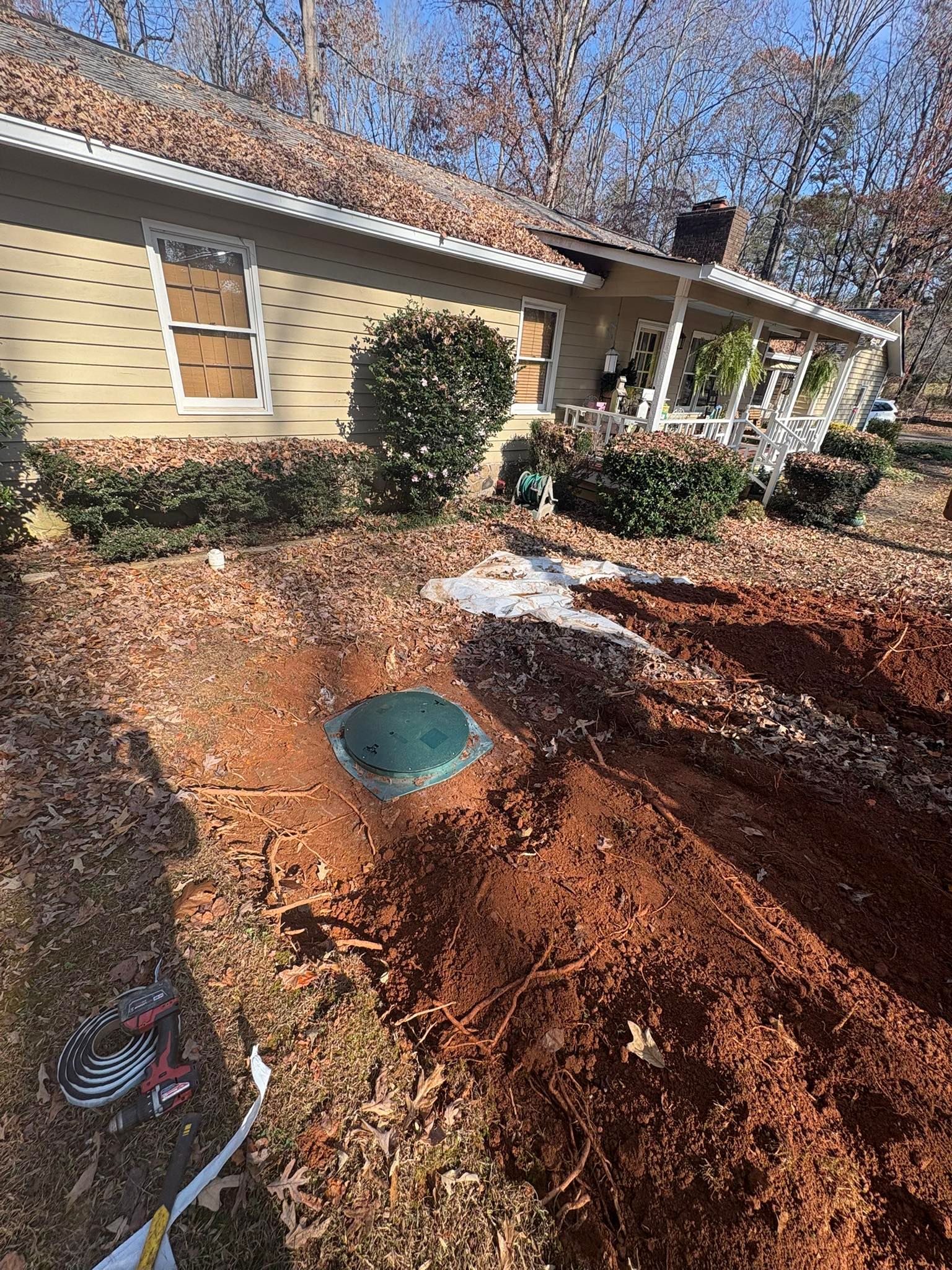 Front yard with dug-up soil around a green utility cover beside a beige house and shrubs