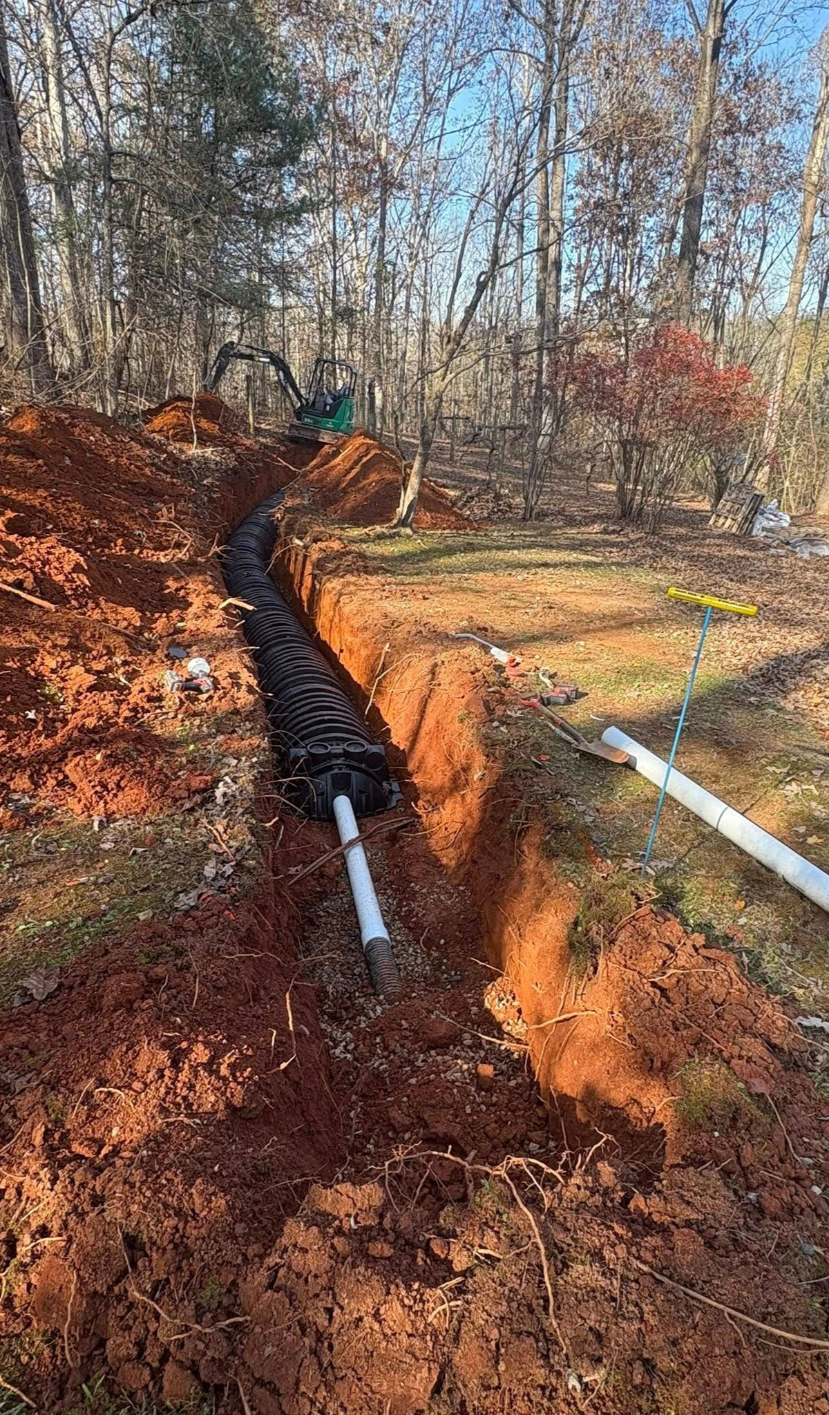 Black pipe laid in a muddy trench on a wooded site, with white PVC pipes nearby.