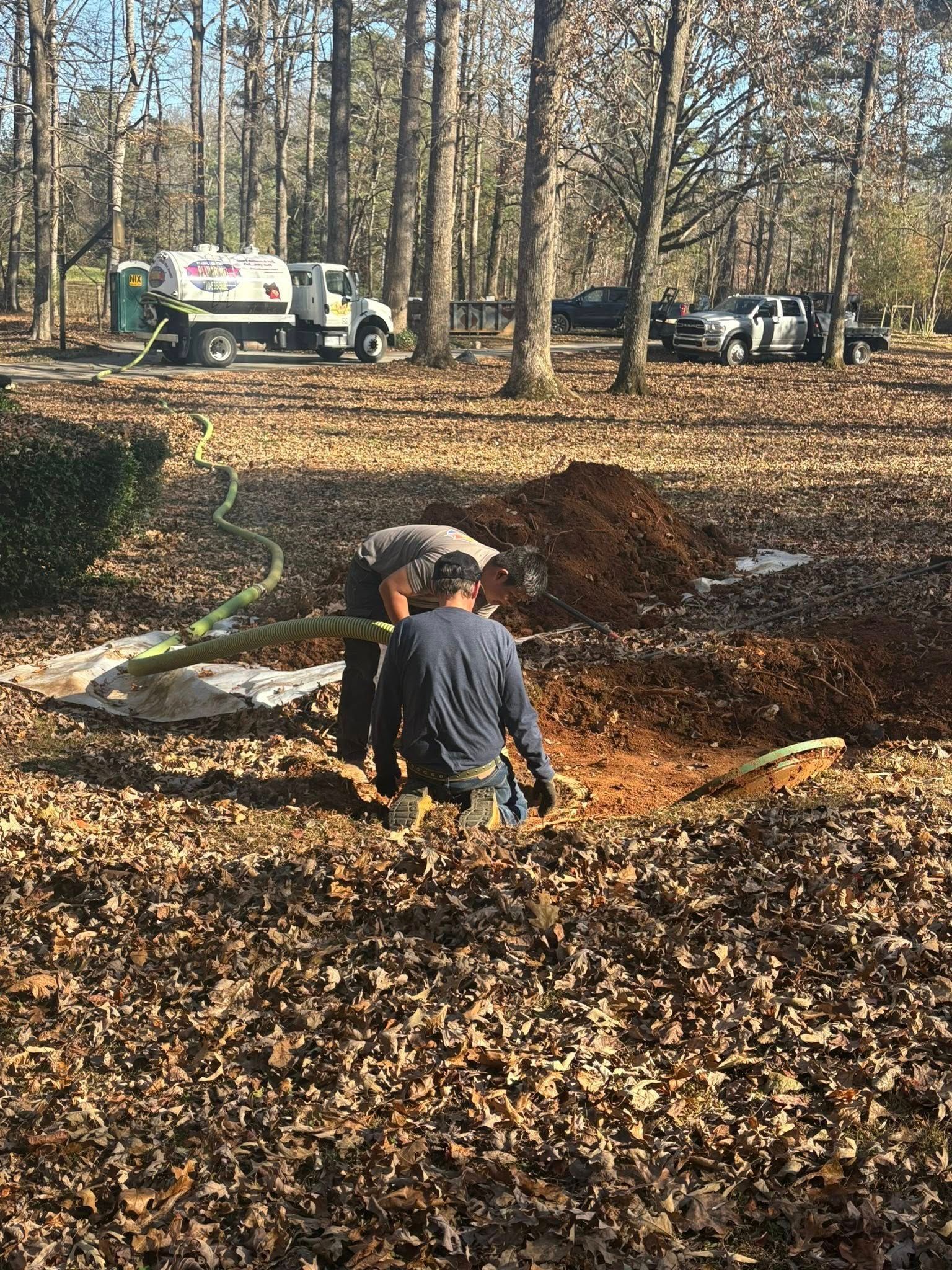 Workers in a wooded park dig a trench near utility trucks and piles of leaves.