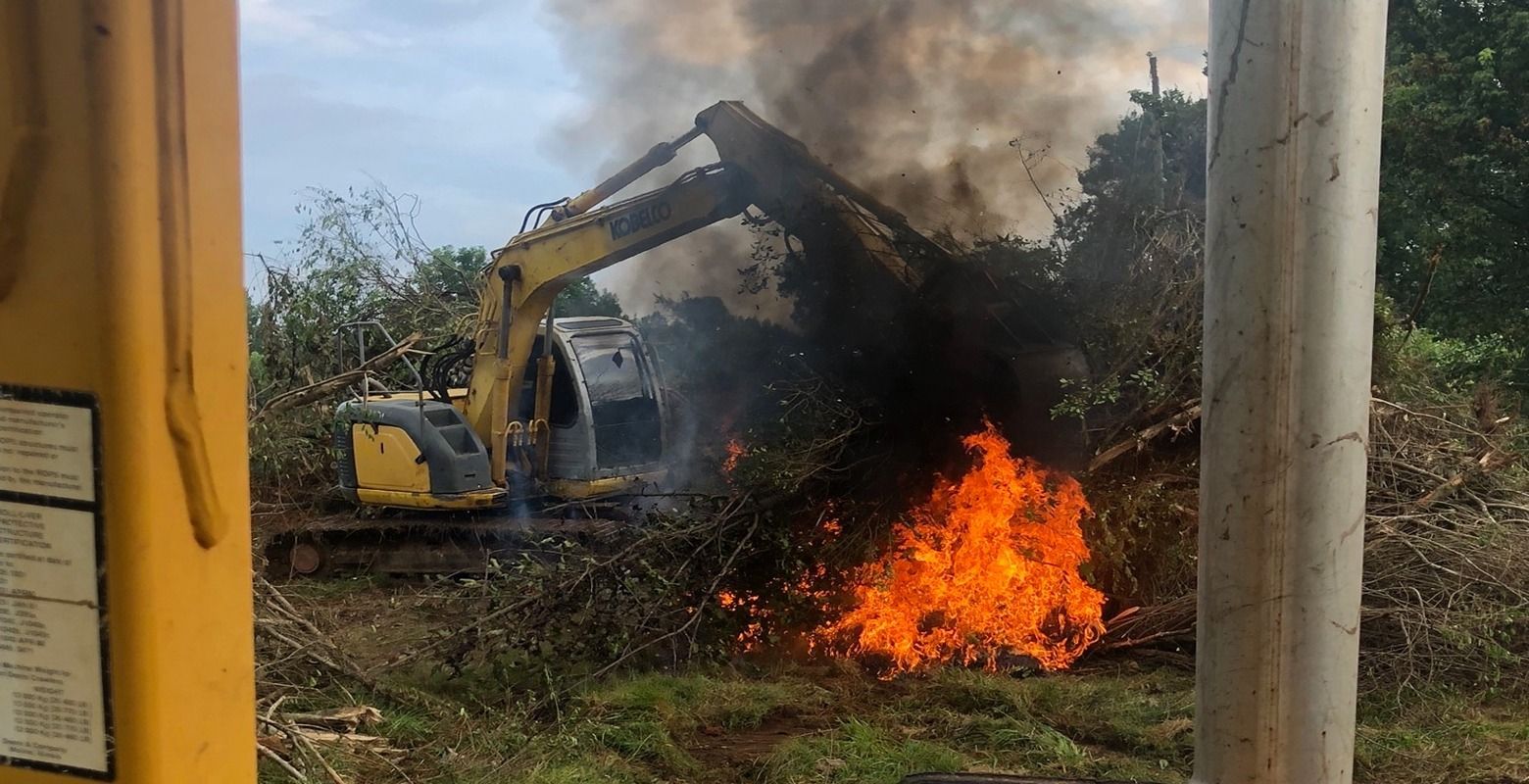 Excavator battles a fire in a brushy area, with thick smoke and flames rising nearby.
