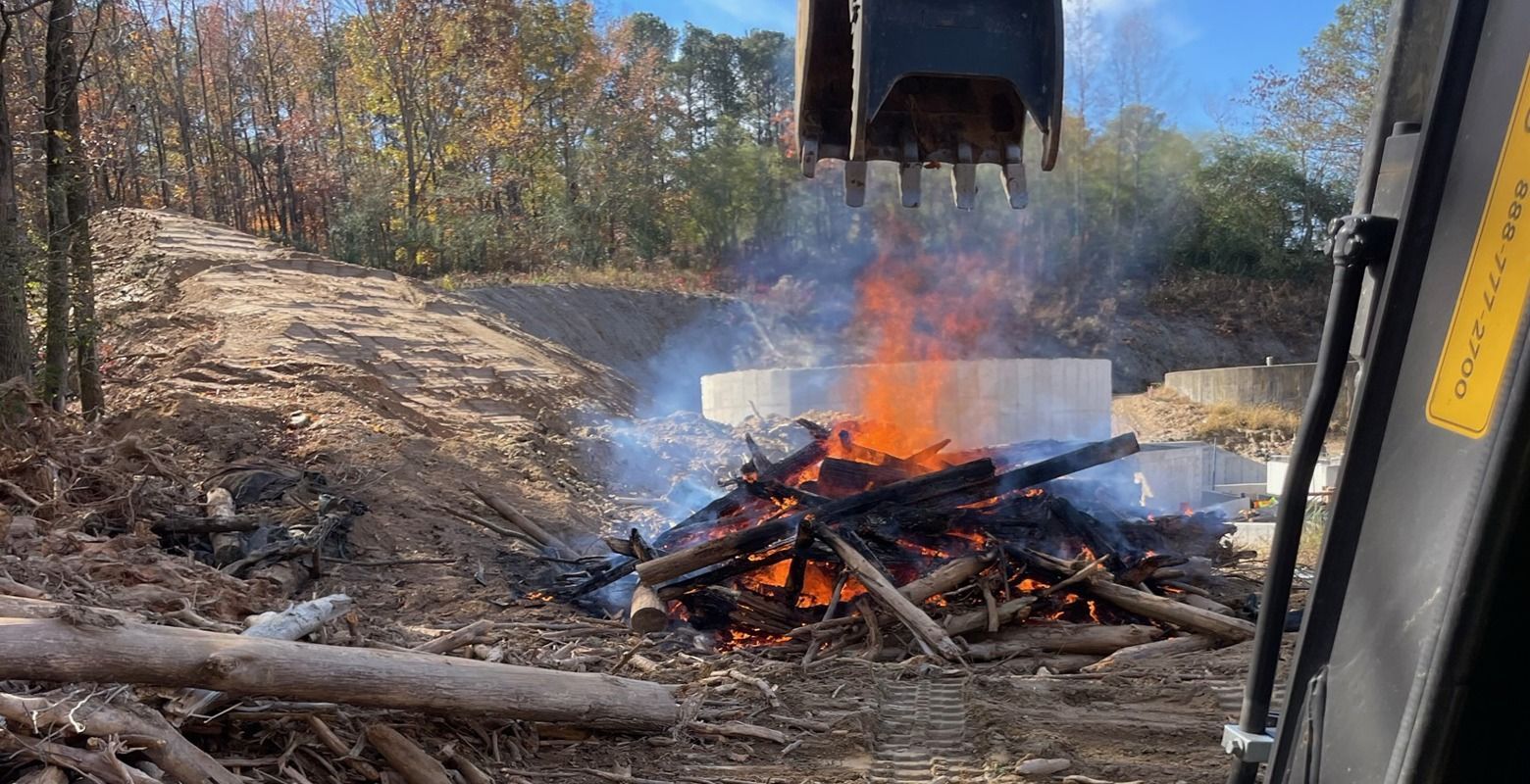 Bulldozer pushes burning debris, with flames and smoke rising in a wooded clearing.