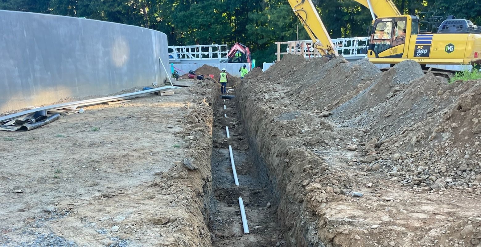 Construction trench beside a wall, with excavators and equipment in the background