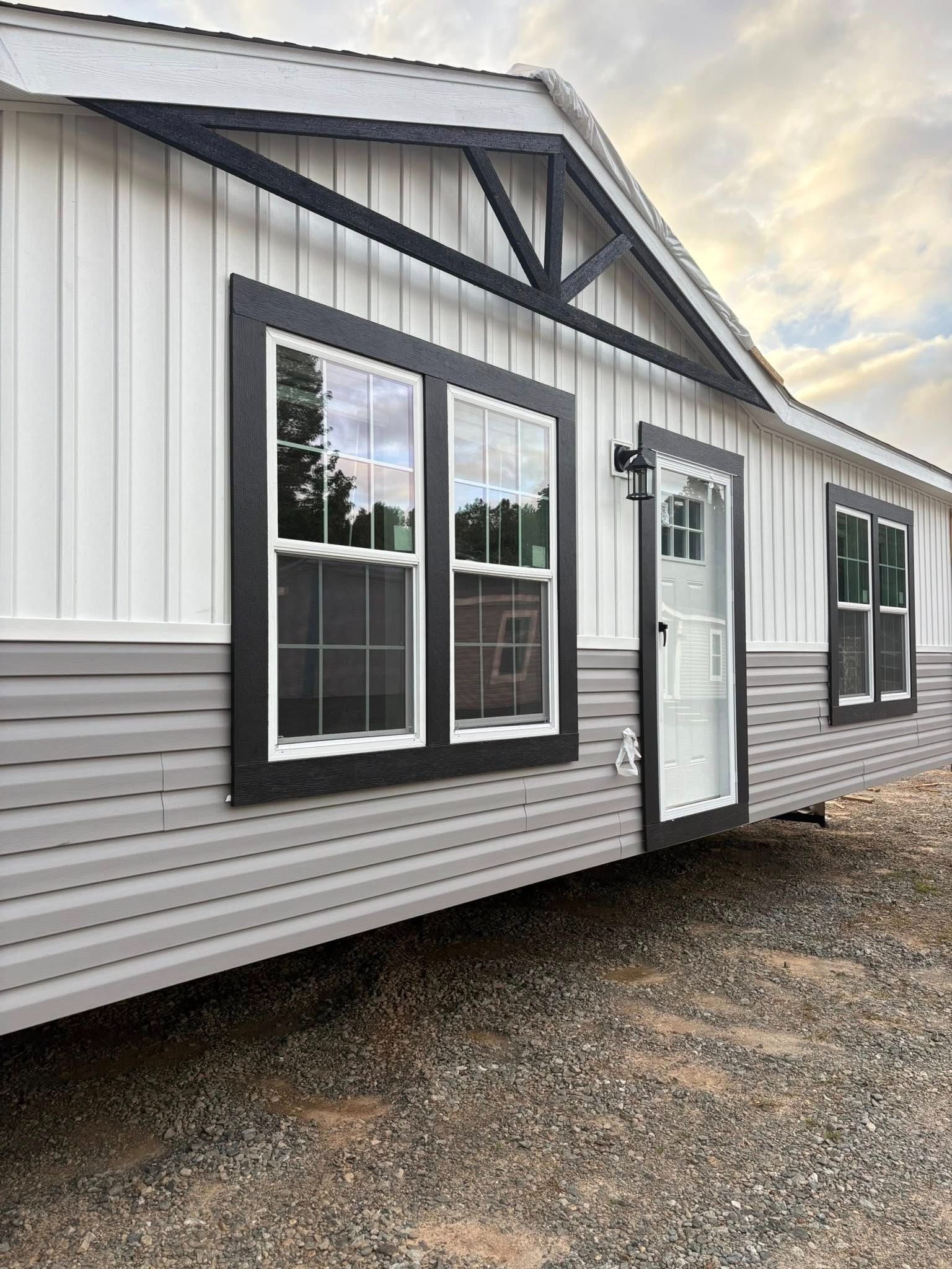 Exterior of a white and gray manufactured home with black-trimmed windows and a front door on gravel.