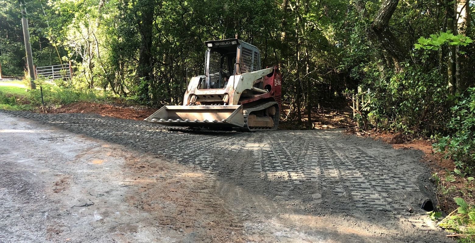 Bulldozer grading a gravel driveway beside a wooded area