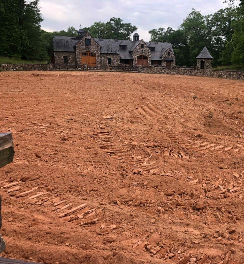 Large cleared dirt lot in front of a stone house and outbuildings, bordered by trees and a stone wall