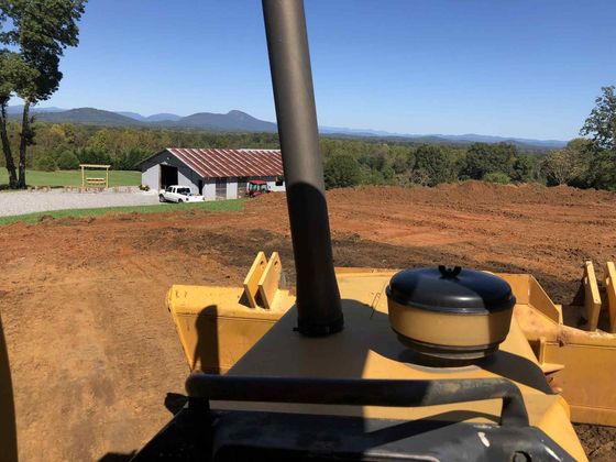 View from a yellow tractor over a dirt field toward a house and mountains under a clear sky