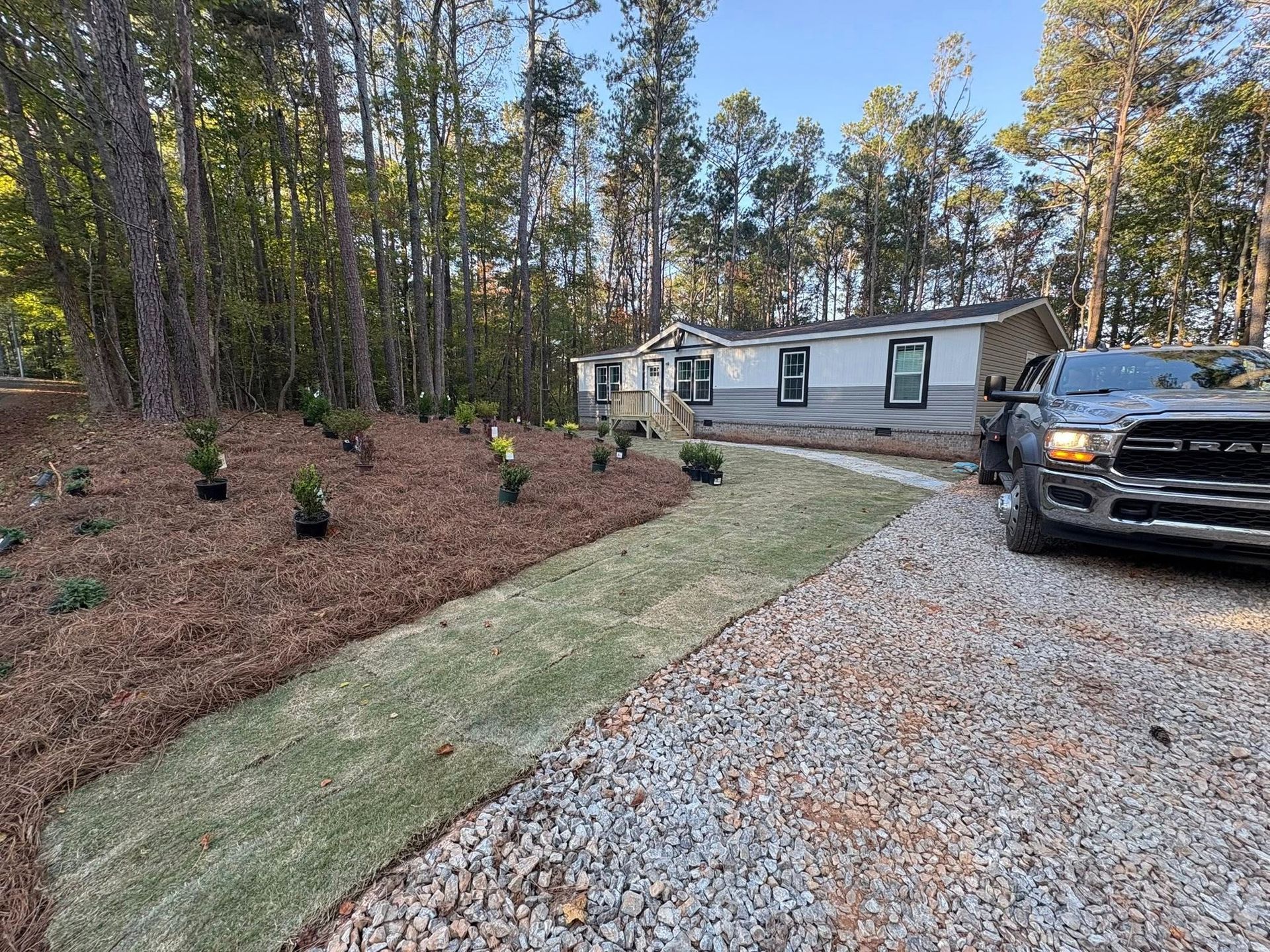 Mobile home with gravel driveway, mulched yard, and parked black pickup beside a wooded lot.