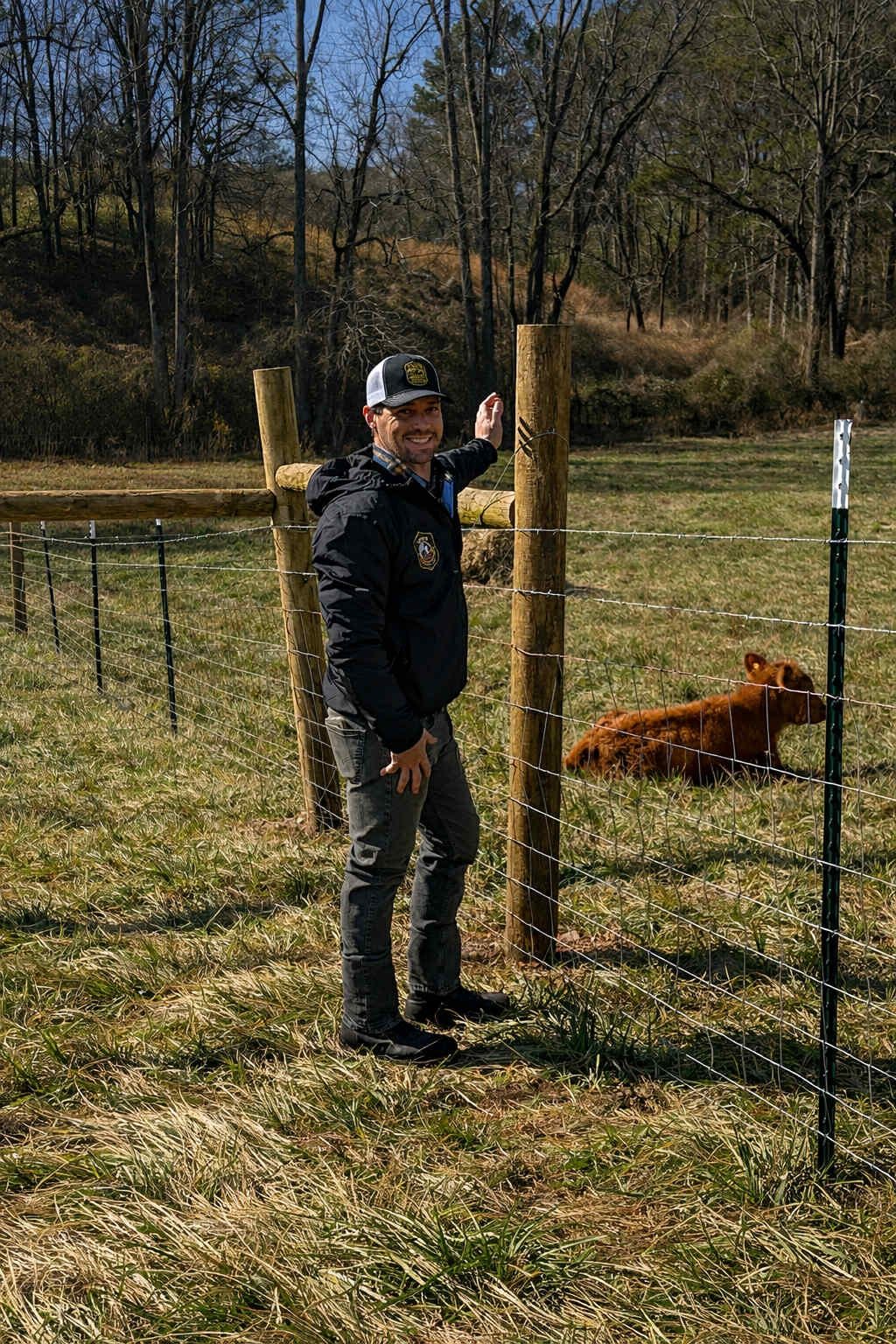 Person in dark clothes stands by a fenced field with a brown dog lying on the grass.