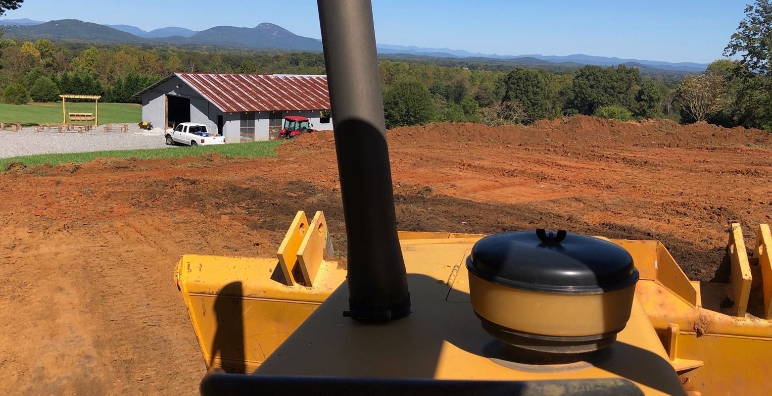 Yellow tractor in a red dirt field with a farmhouse and hills in the distance