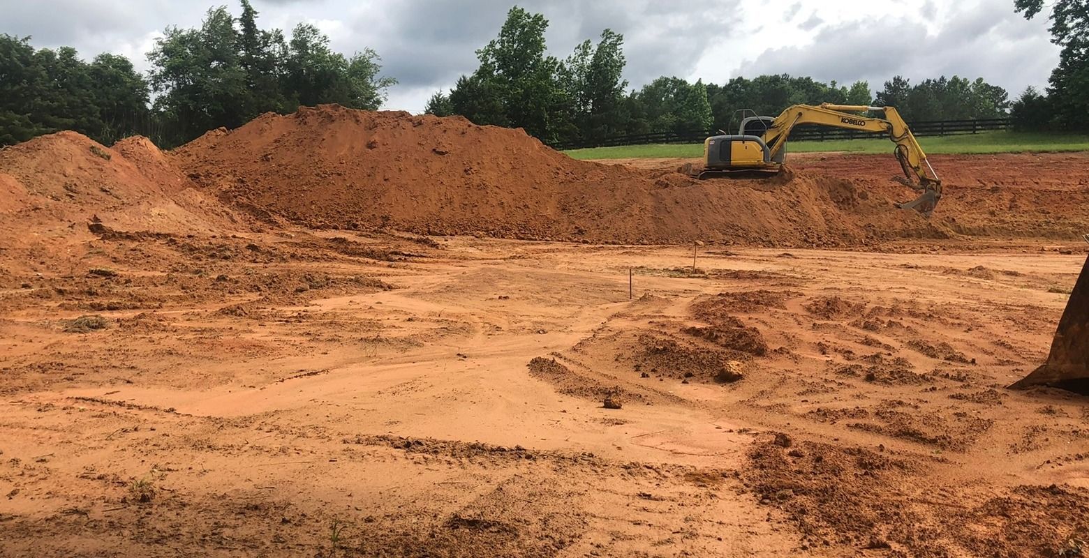 Yellow excavator at a muddy construction site with large dirt piles and trees in the background