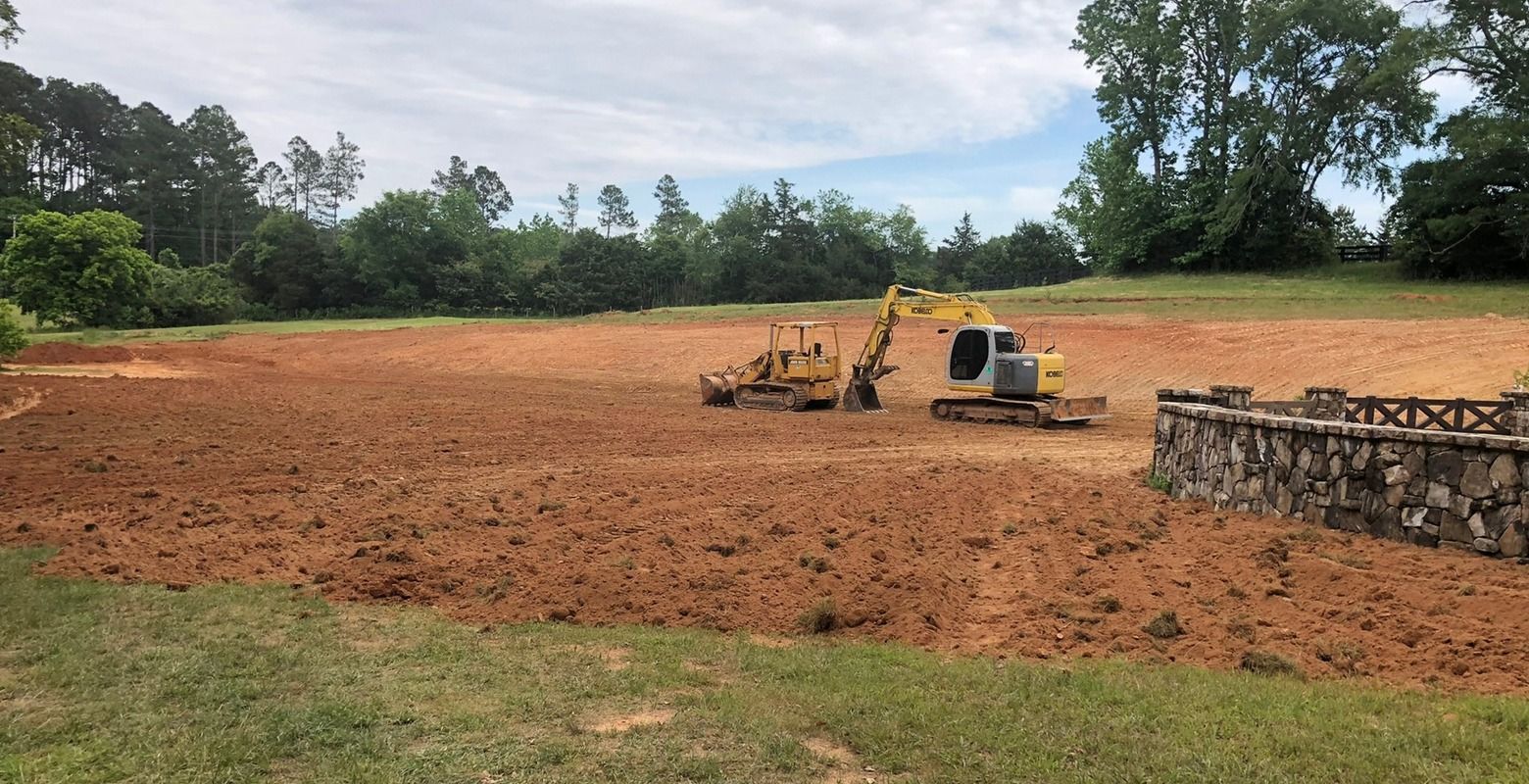 Small yellow excavator leveling a reddish dirt field beside a stone wall, with trees in the background