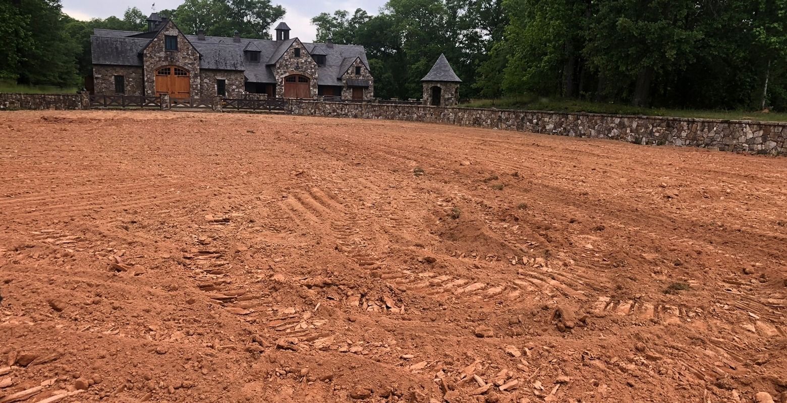Plowed reddish-brown field with a stone house and trees in the background