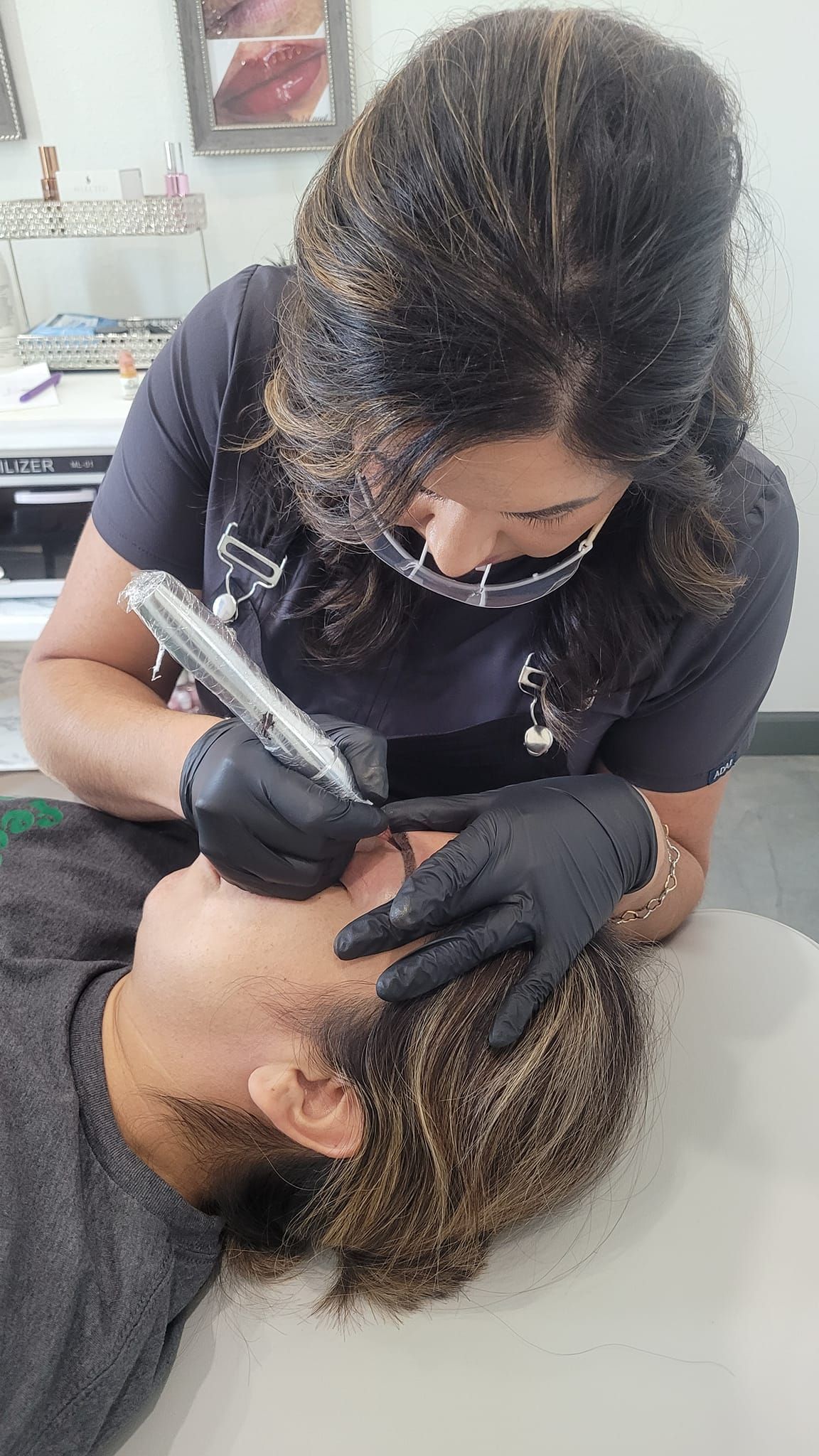 Woman with black gloves performs eyebrow microblading on a person lying on a table.