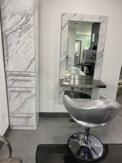 A hair salon station with a marble-look storage cabinet, mirror, stylist chair, and stainless steel table.