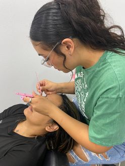 Woman applies eyelash extensions to a client. The stylist wears glasses and a green shirt. They're in a salon.