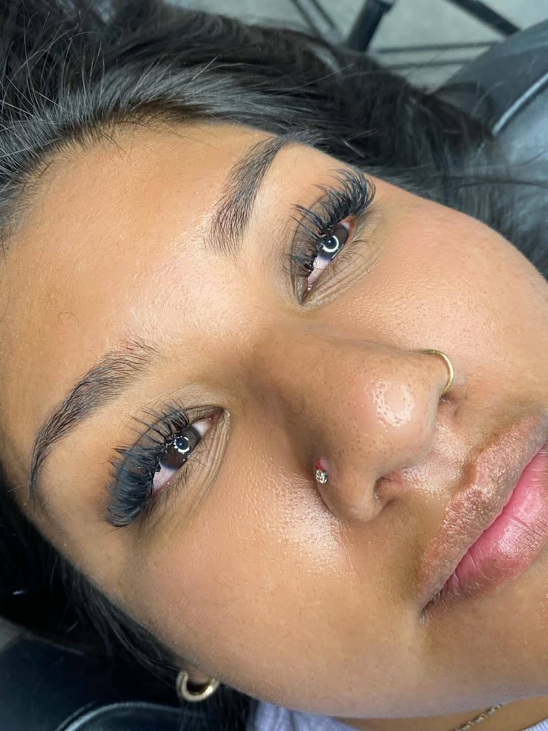 Woman with dark skin, filled-in eyebrows, long eyelashes, and nose piercings, in close-up.