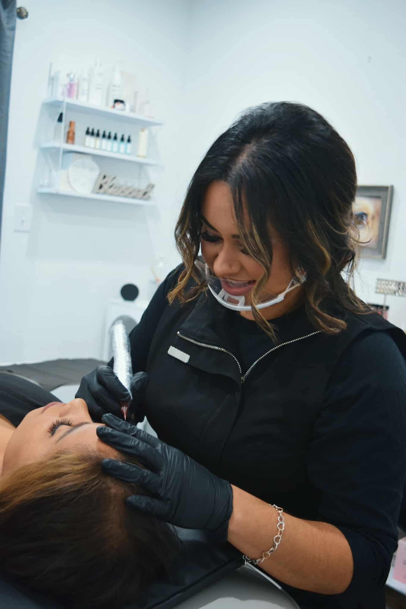 Woman in black gloves and vest, performing eyebrow microblading on a client in a salon.