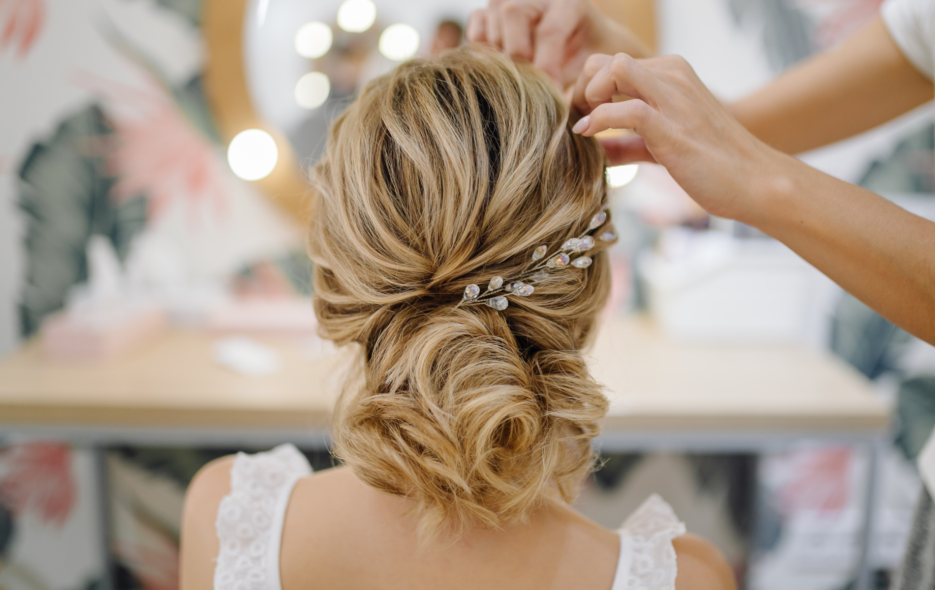 A woman has her hair styled in a bridal updo, with a stylist adjusting a decorative hairpiece.