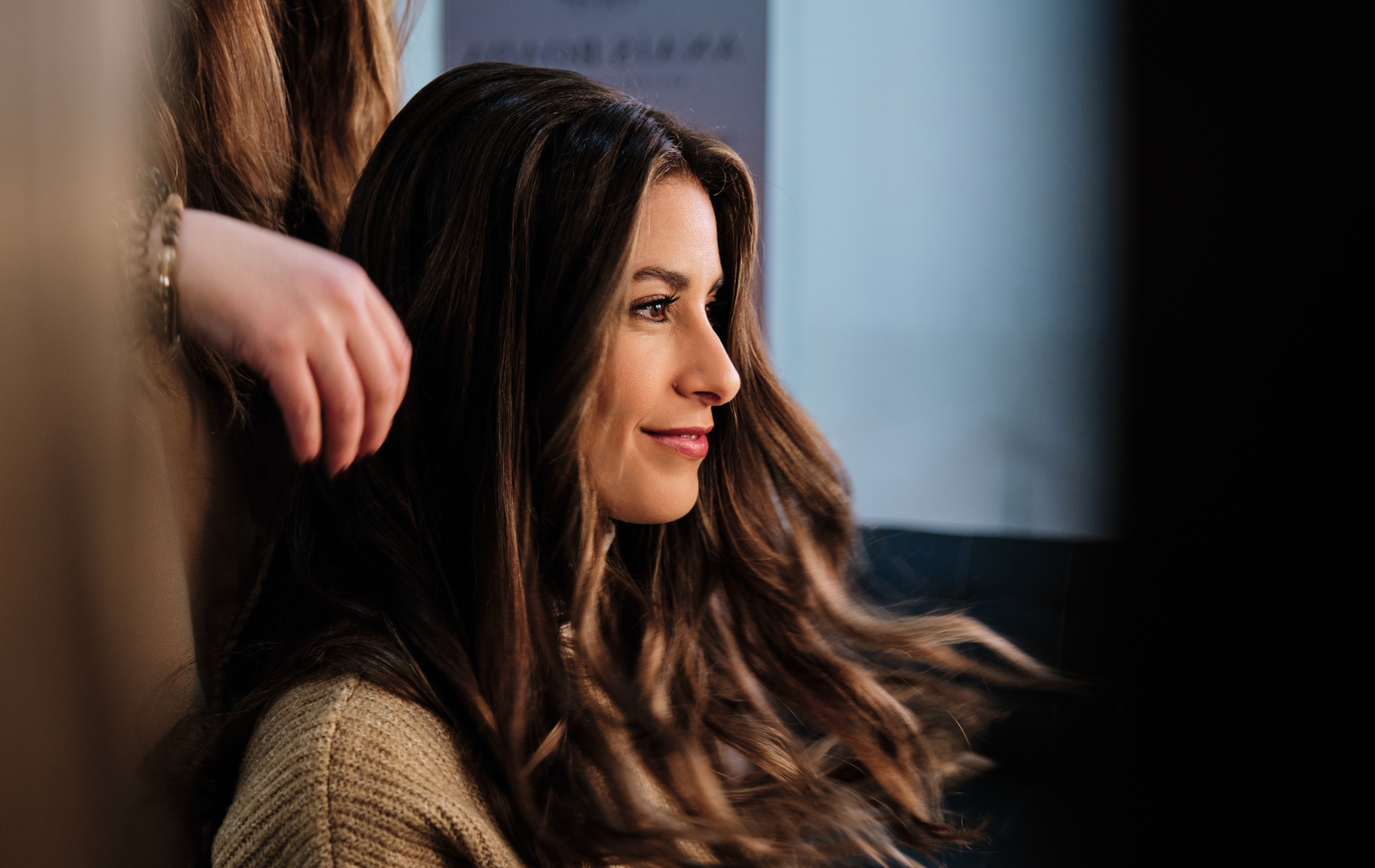 Woman with long, wavy brown hair smiles while someone adjusts her hair in a salon.