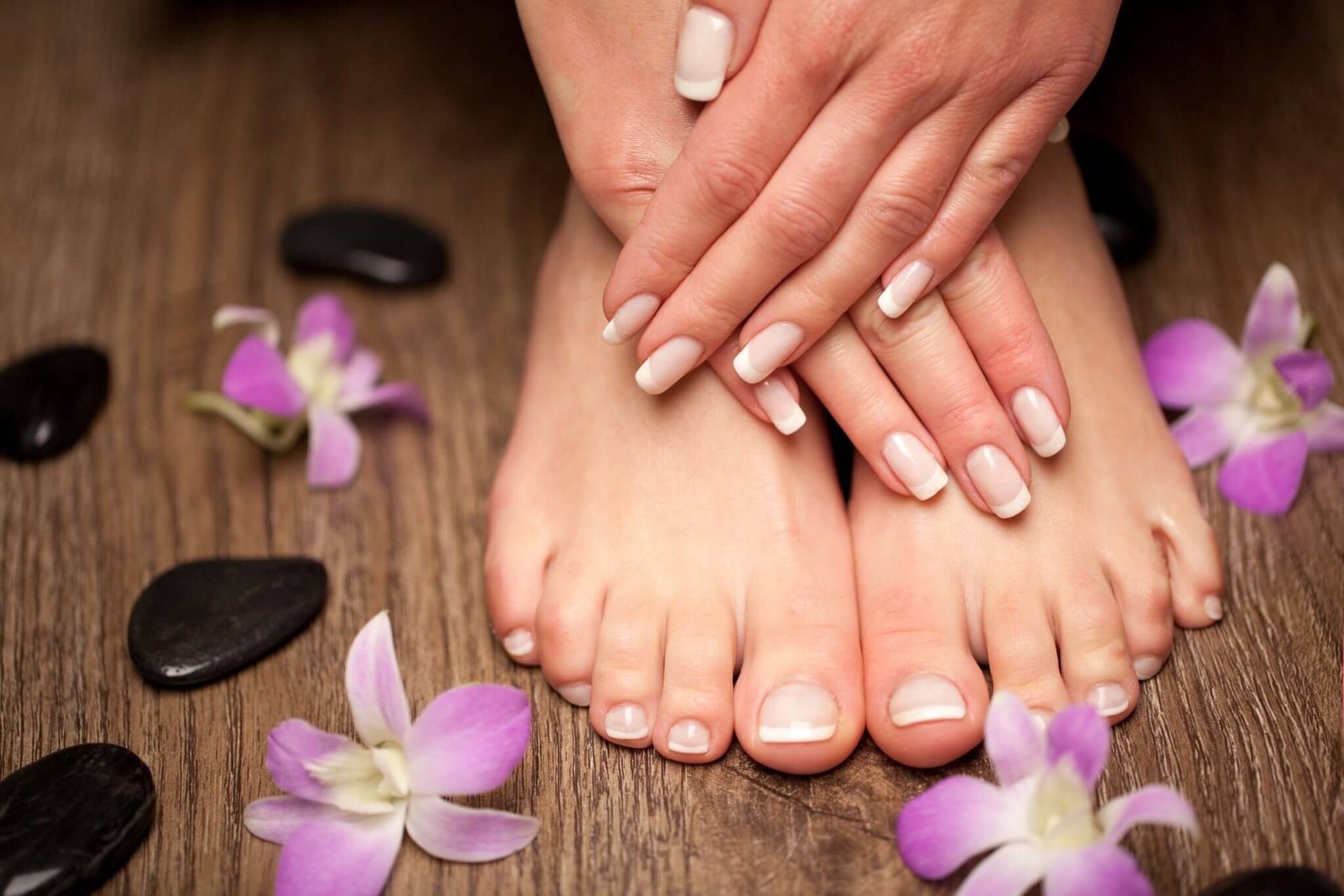 Hands with manicured nails rest on feet with pedicured toes, surrounded by flowers and stones.