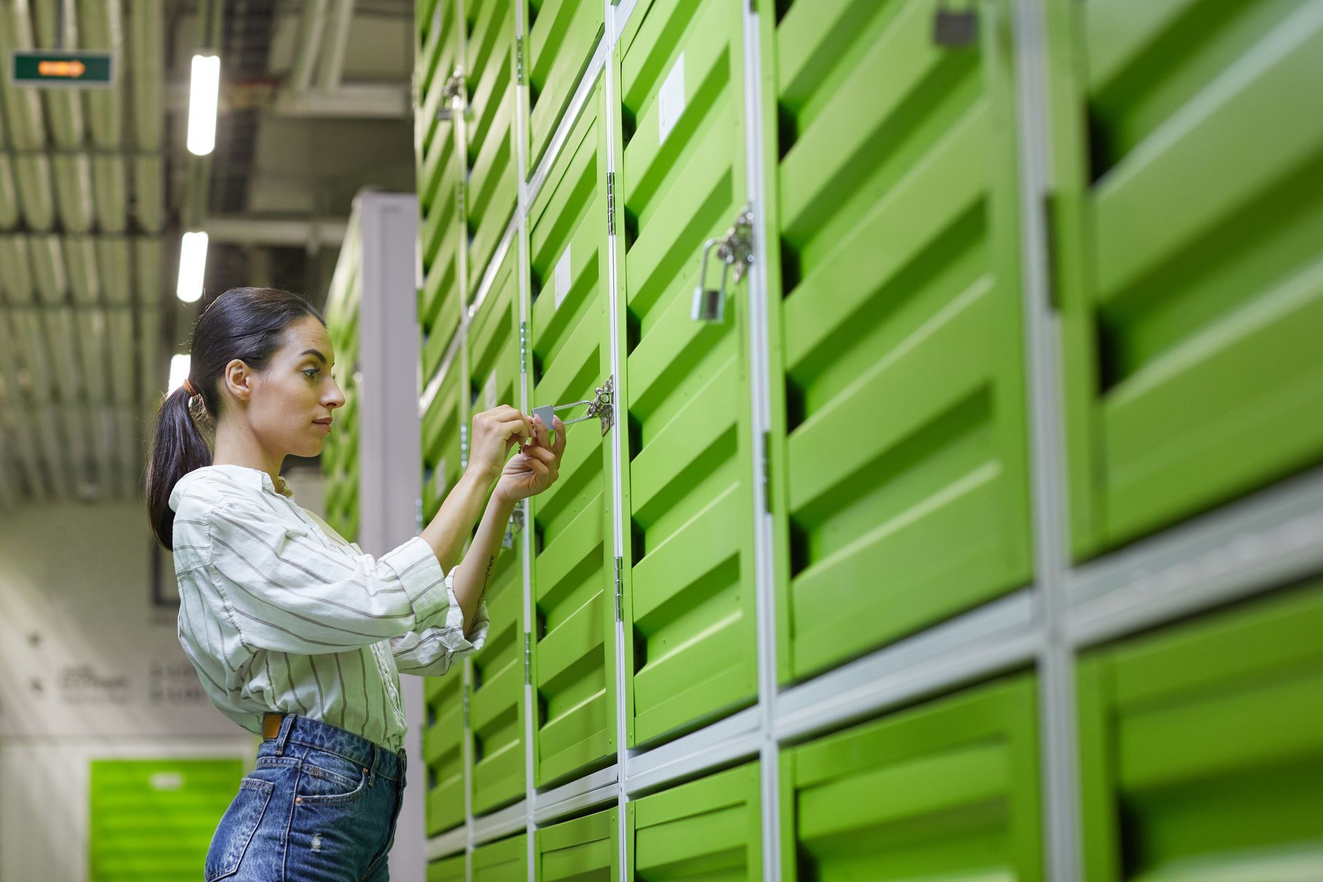A woman wearing casual clothing opens a green storage unit, unlocking the padlock.