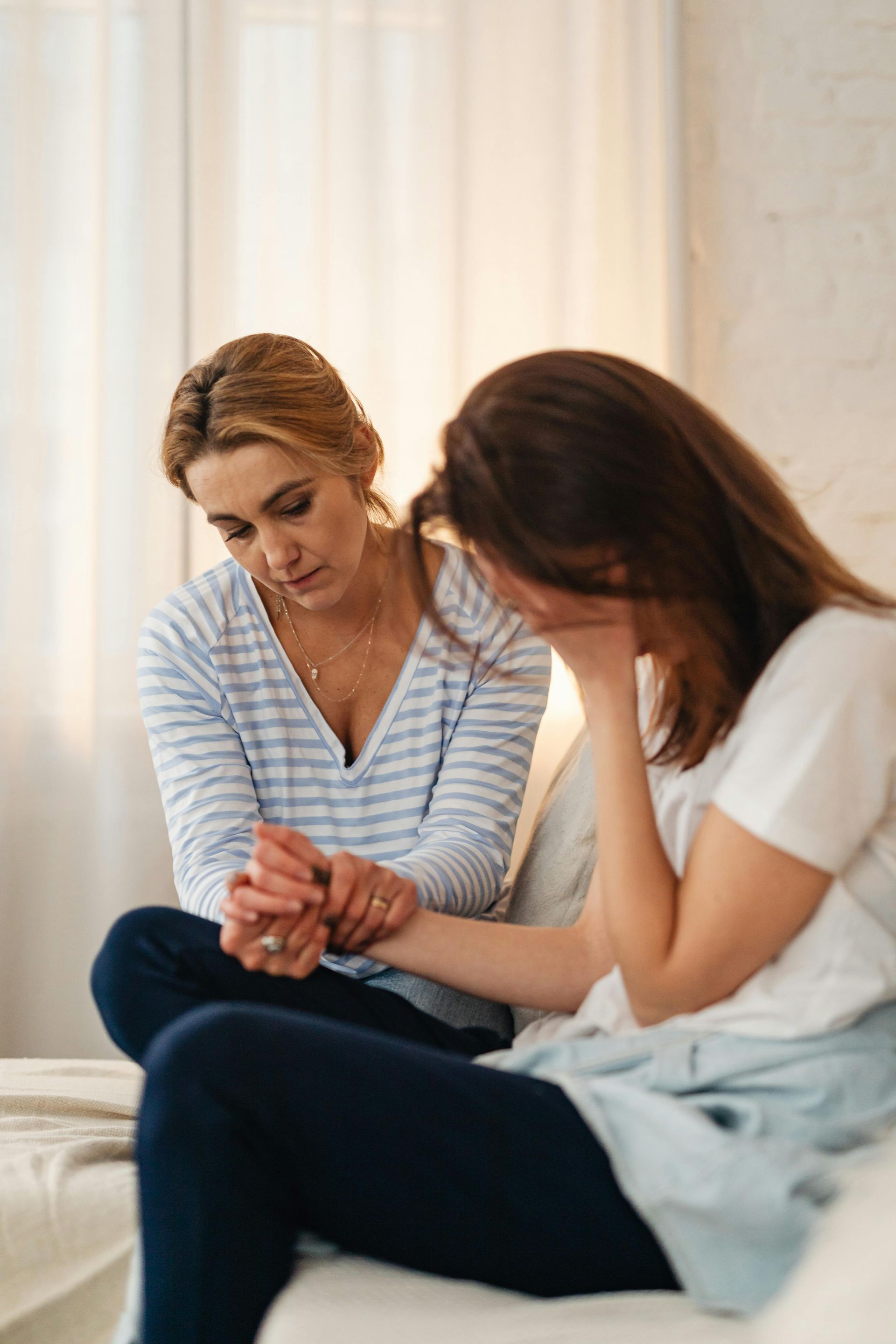 Woman comforts another, holding her hand and offering support. Light-filled room.