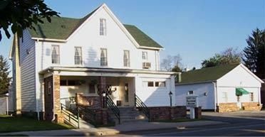 White, three-story building with green roof, brick porch, ramp, and attached garage. Sunny day.
