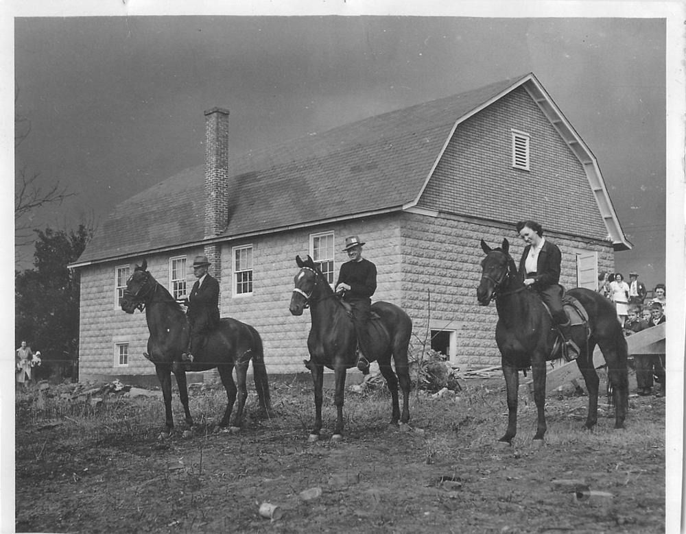 Three people on horseback in front of a stone barn.
