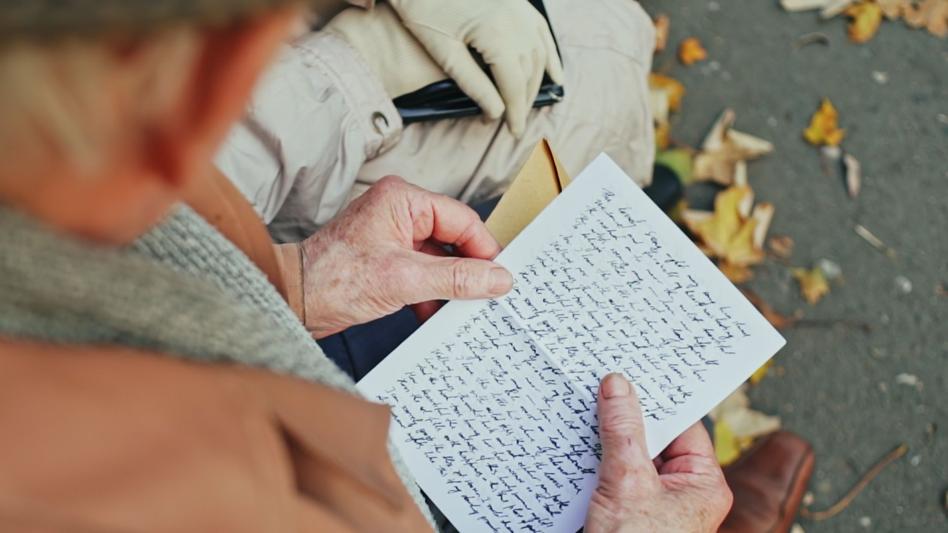 Elderly person reading a handwritten letter outdoors, holding the paper with wrinkled hands, fall leaves nearby.