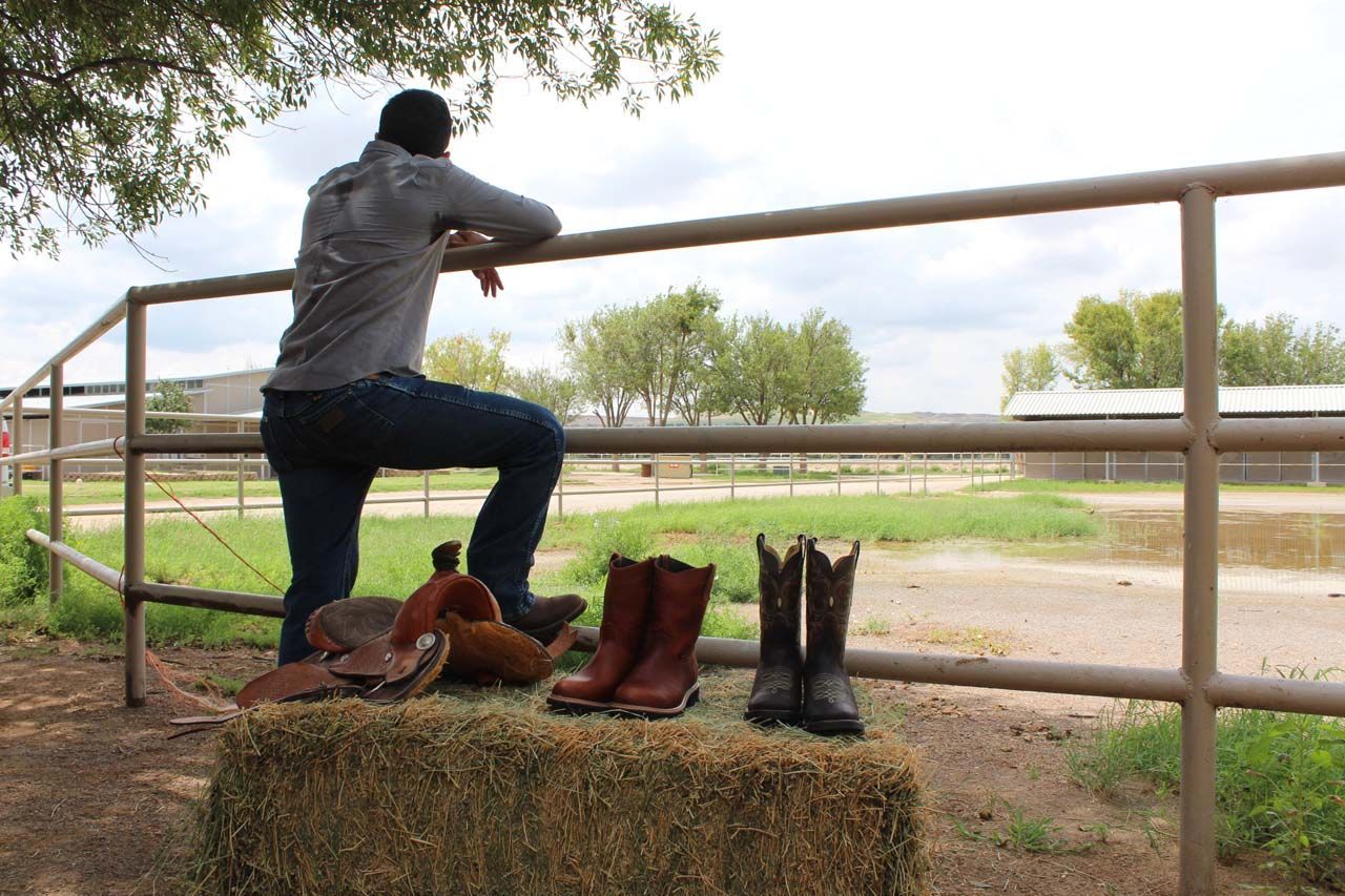 A man leaning on a fence with cowboy boots on a bale of hay.