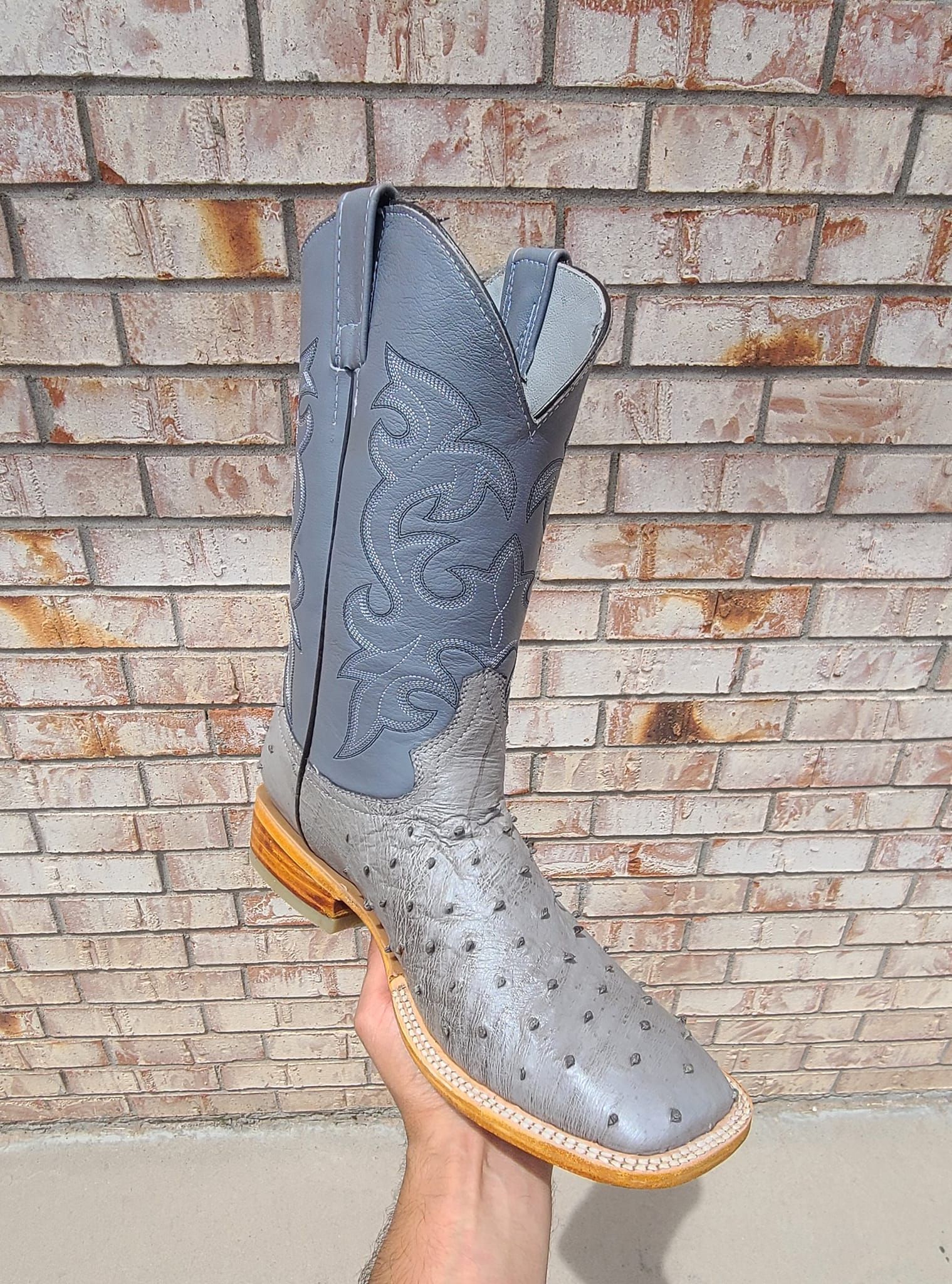 A person is holding a pair of cowboy boots in front of a brick wall.