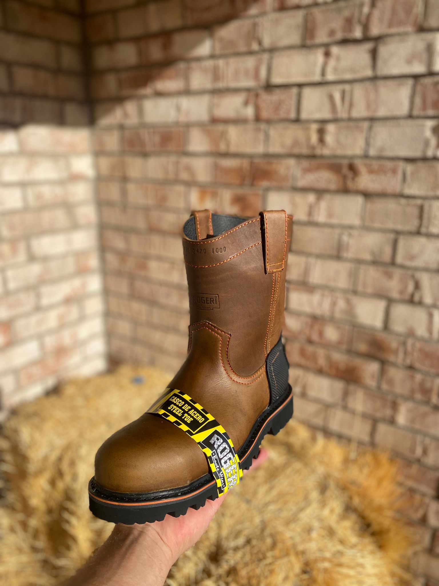 A person is holding a pair of brown cowboy boots in front of a brick wall.